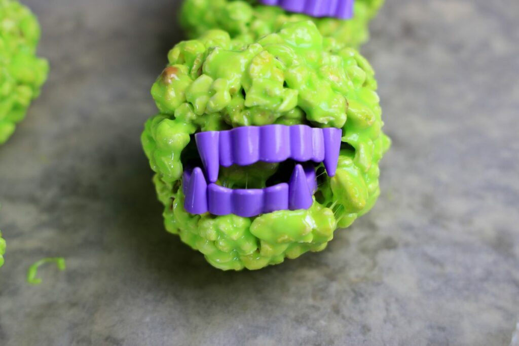 Green popcorn ball with purple vampire teeth on parchment paper.