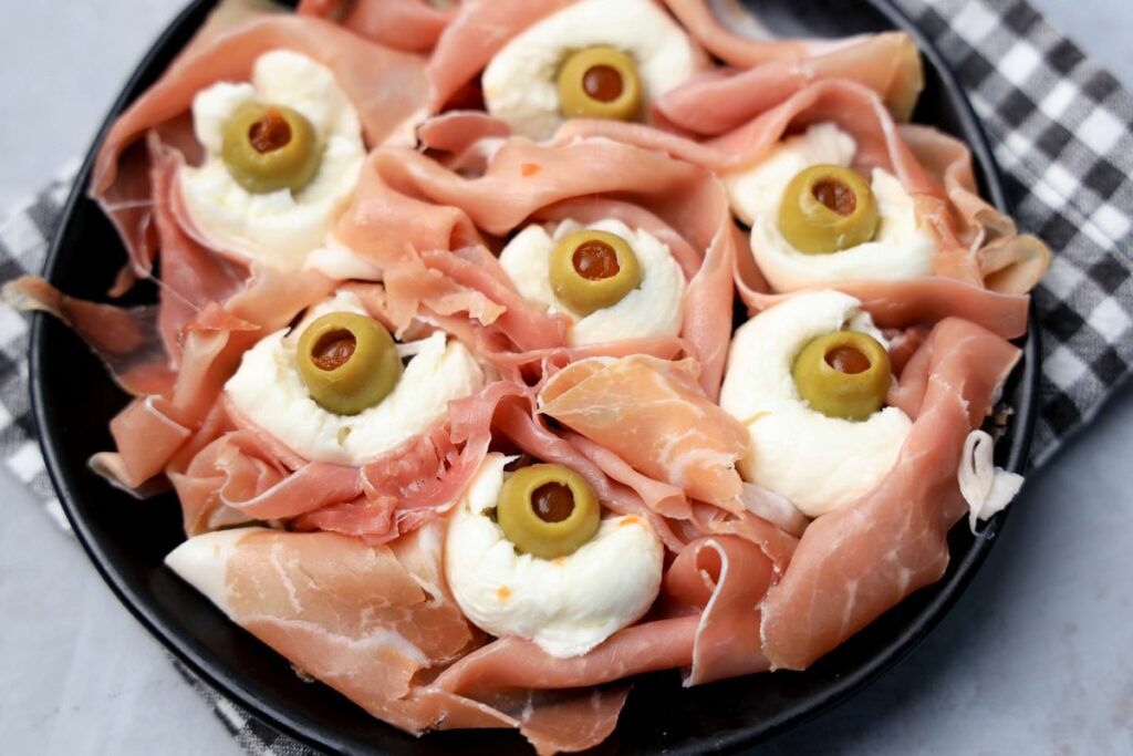 Fresh mozzarella, green olives, and prosciutto to make spooky eyeballs on a black plate.